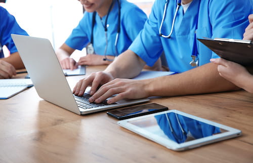 A group of med students in scrubs work on tablets and laptops at a desk. Shown from the shoulders down.