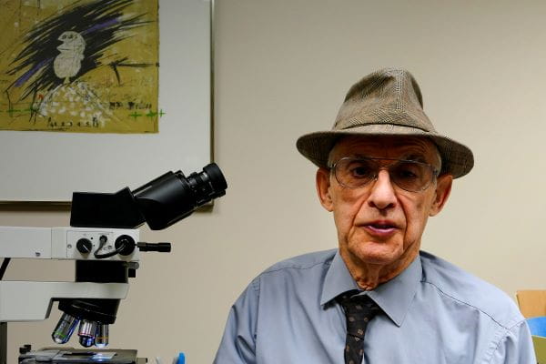 Photo of Dr. Larry Roth, wearing a fedora, looking at the camera while sitting next to an laboratory instrument