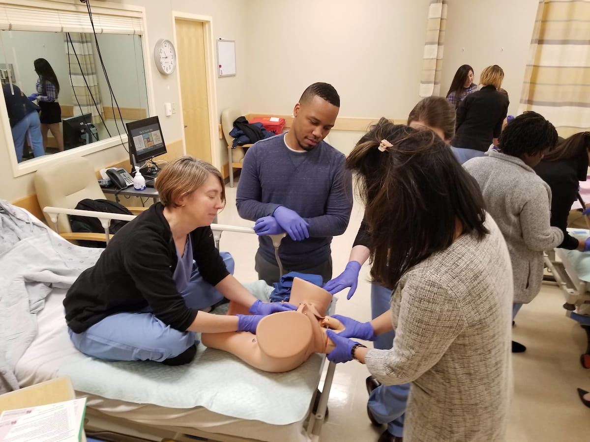 Three trainees work on a manikin while a female faculty member supervises, sitting on hospital bed in simulation center