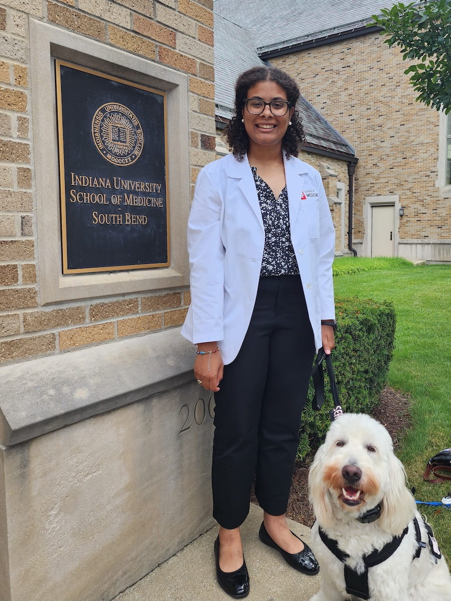 Kennedy Onuoha wearing her white coat and standing with her goldendoodle Aura by the IU School of Medicine--South Bend sign outside a campus building
