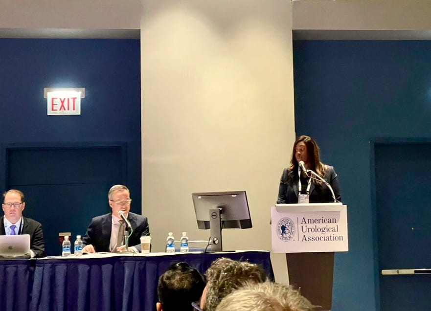 Chinade Roper in business attire at podium, presenting research to the American Urological Association while two men in suits listen from table at front of room