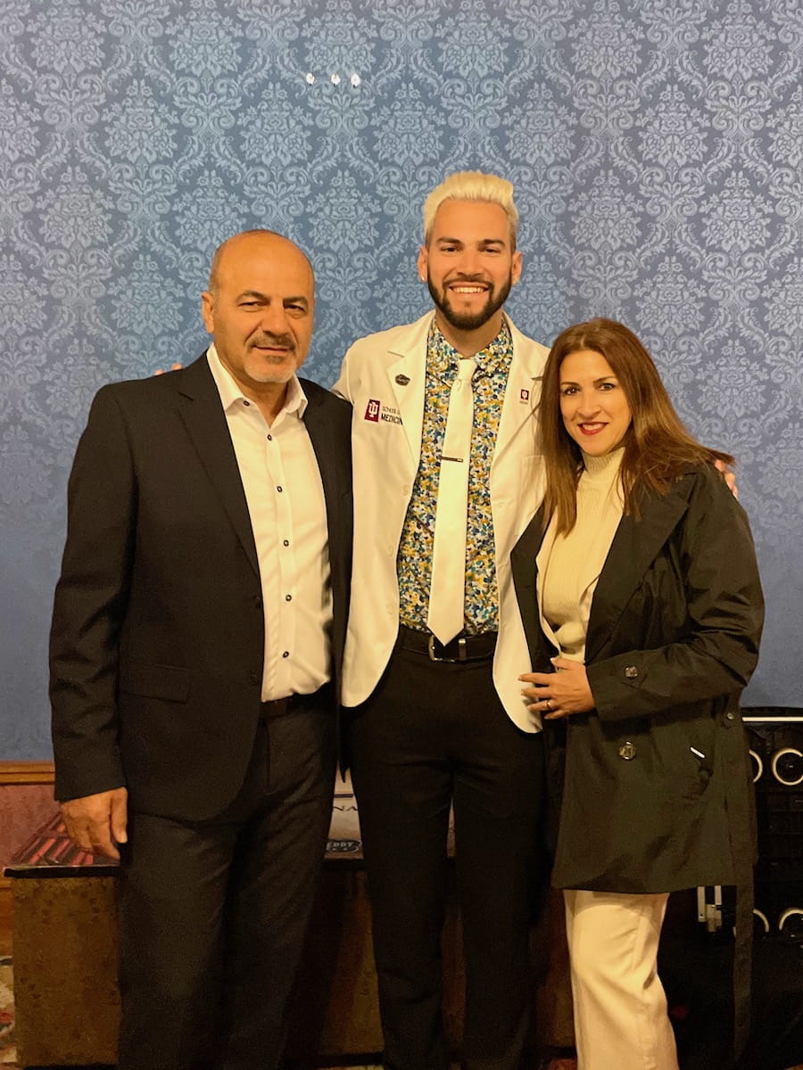 Devin Gantzios-Cros stands with arms around his father and mother, wearing his white coat after the Class of 2024 White Coat Ceremony