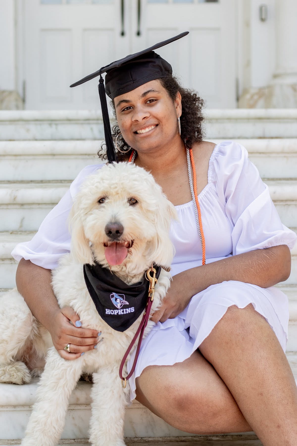 Kennedy Onuoha in graduation cap sitting on steps with goldendoodle Aura, wearing a black Johns Hopkins bandana.