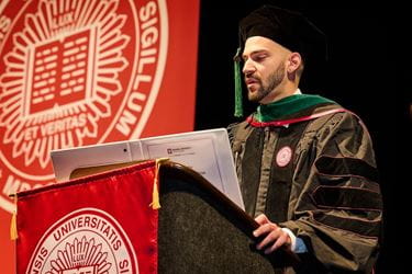 Devin Gantzios-Cros in his cap and gown standing and speaking at podium with IU School of Medicine logo flags, at graduation 2024