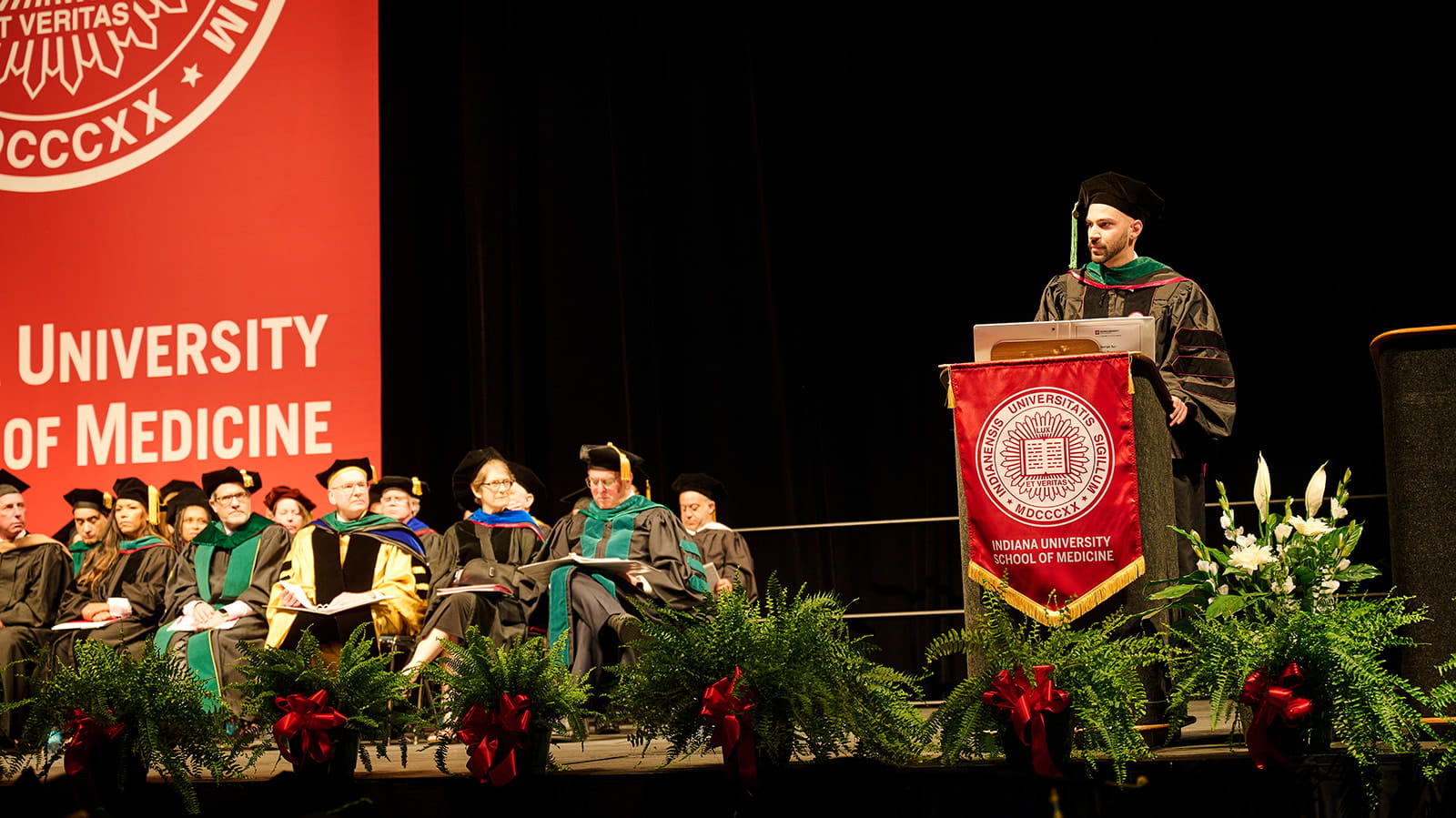 Devin Gantzios-Cros on stage with platform party as he speaks, wearing his cap and gown at the Graduation 2024 ceremony.