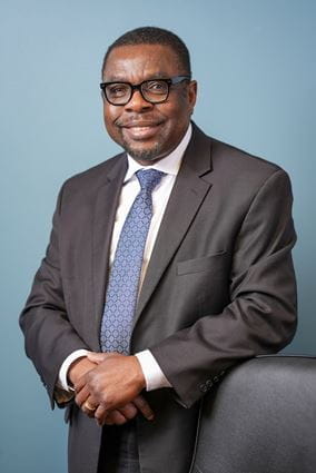 Kola Okuyemi in suit and tie leaning on chair, with blue background