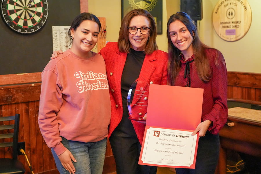 Maria Del Rio-Hoover standing in center wearing red jacket and holding her award next to students Laura Cordero and Olwen Menez, holding a certificate