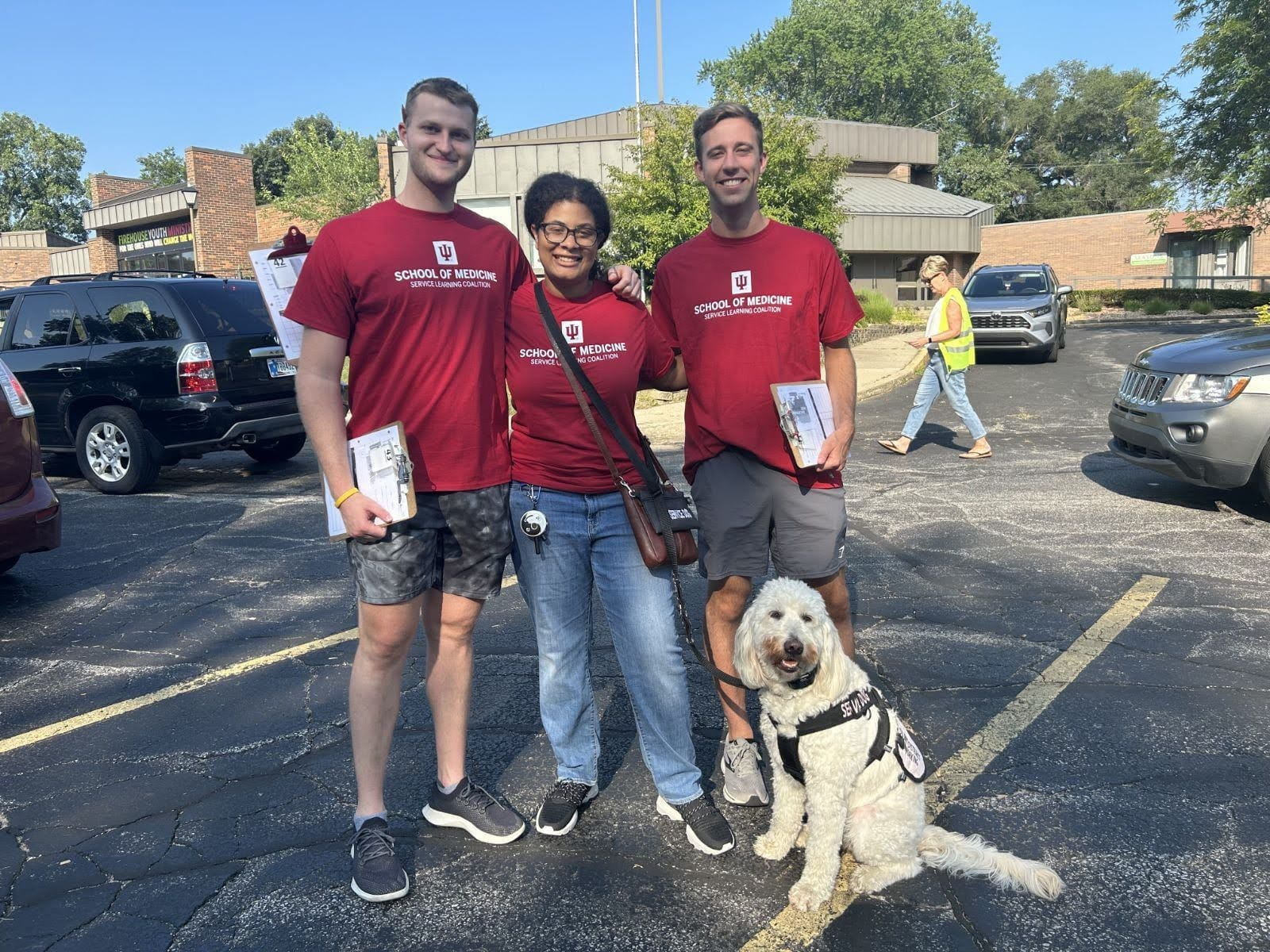 Kennedy Onuoha and two male classmates in matching IU School of Medicine Tshirts stand in a parking lot with Aura the goldendoodle service dog after volunteering at a community food pantry.