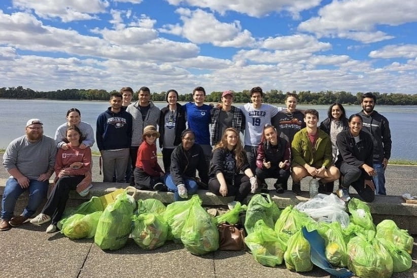 a large group of students stand with their arms around each other in front of a large river. in front of them are 25+ bags filled with trash. There is a blue sky with scatter clouds overhead.