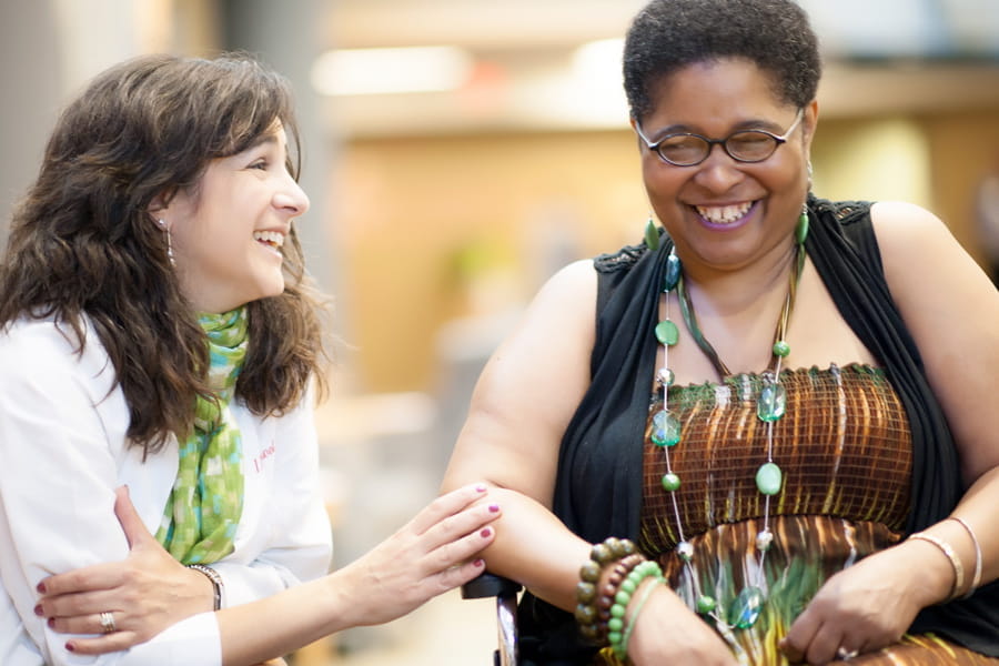 a physician laughs and lightly places a hand on the arm of a patient in a wheelchair who is smiling