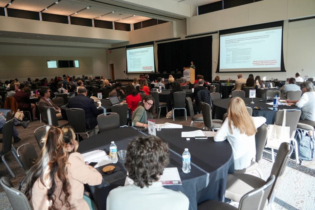 a room full of people sitting at tables while a speaker stands at the podium on the far side of the room