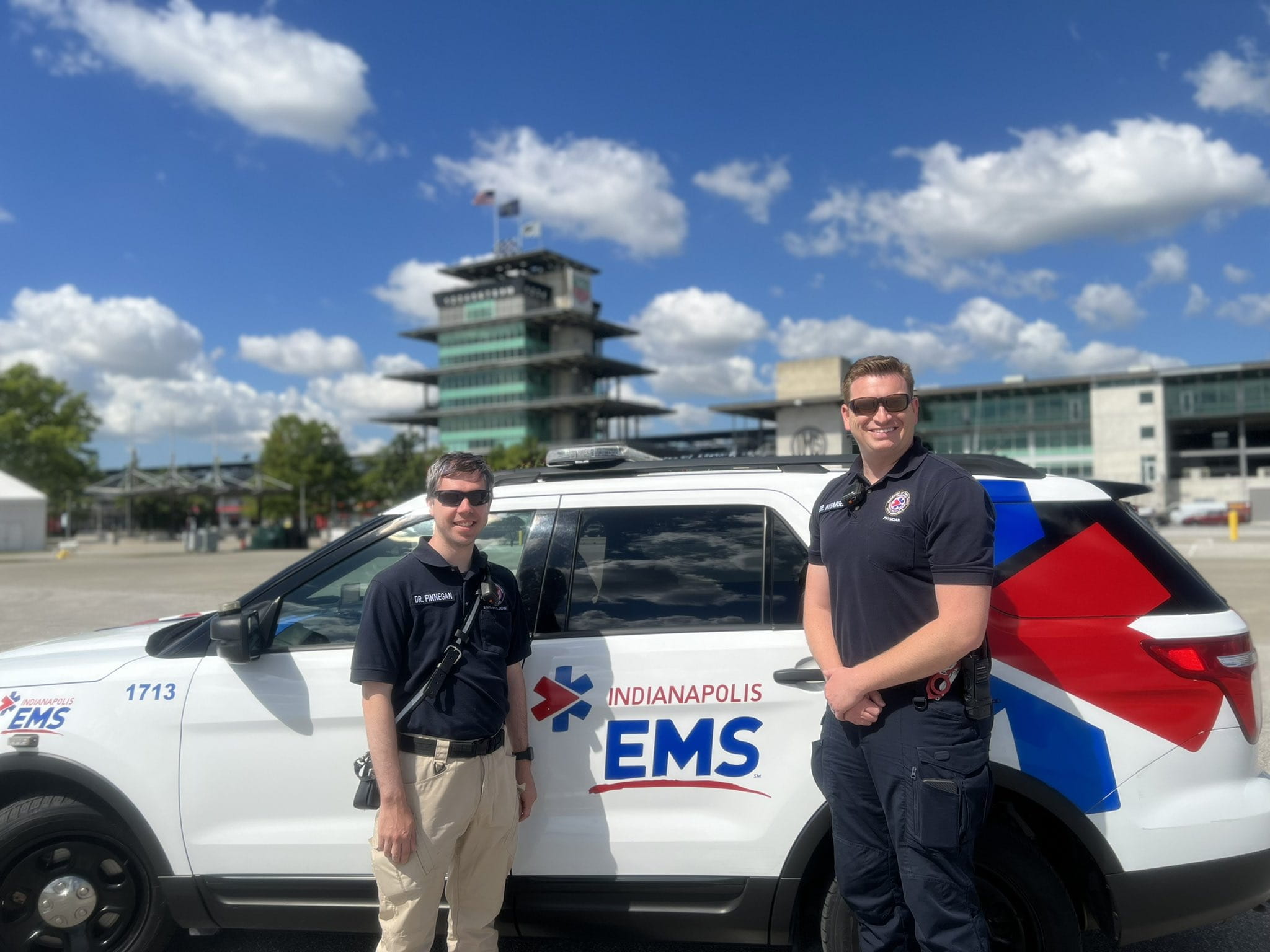 Two doctors smile in front an EMS vehicle on a sunny day. 