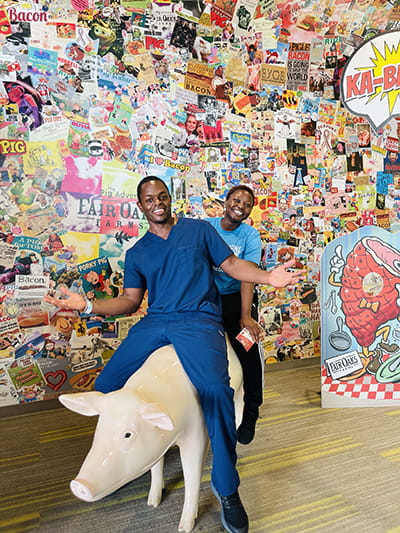 two residents smile as they sit on the back of a statue of a pig, in front of a wall covered in cartoons