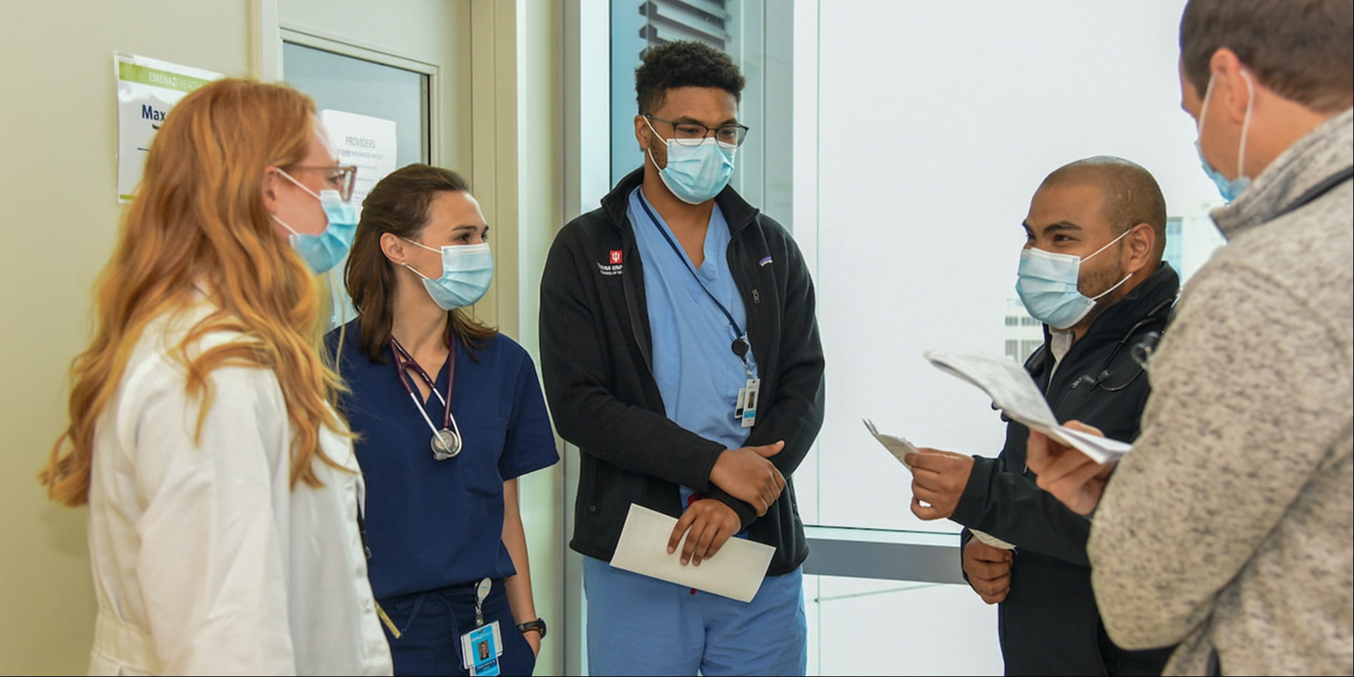 A mixed-gender group of young physicians in face masks consult each other in a brightly-lit hospital hallway.