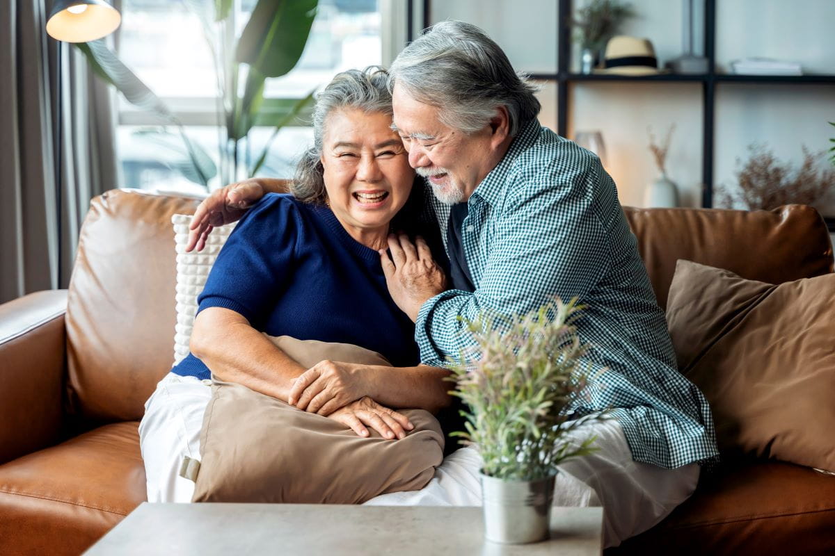 an older asian couple sits together on their couch
