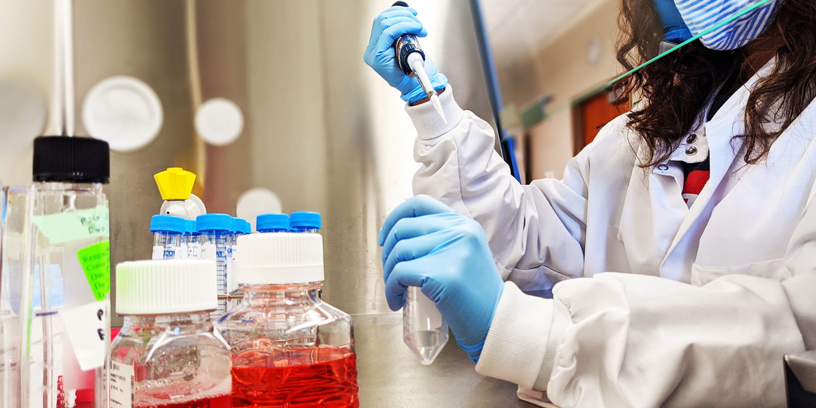 a scientist with long curly hair wearing a mask and lab coat prepares samples in the genetics testing laboratory
