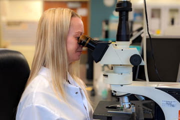 a lab member with straight long blond hair wearing a white lab coat looks into a microscope