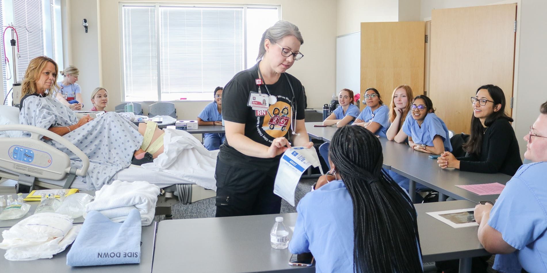 A nurse educator shows a resident intern the blood pressure cuff. Other interns watch from the side. A simulated patient sits on the hospital bed in the classroom.