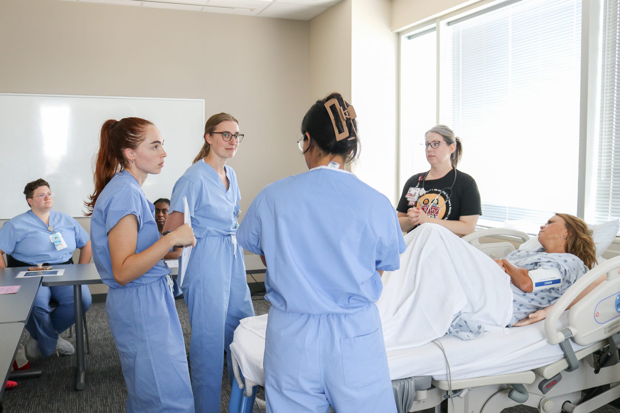 Three resident interns stand around a hospital bed in the simulation classroom. A patient lies in the bed with a nurse educator beside her. The residents discuss the appropriate care for the patient.