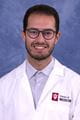 Muhammad wearing a white lab coat and glasses, with short dark hair and beard.