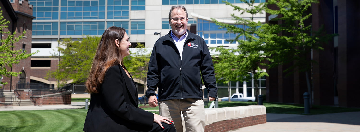 A man and woman are engaged in a friendly conversation outdoors on a sunny day in front of a university building. The man is wearing a black jacket with the logo 'School of Medicine, Pathology,' while the woman, seated on a low wall, listens attentively. The background features green trees, a lawn, and a multi-story building with large windows, creating a professional yet welcoming campus environment.