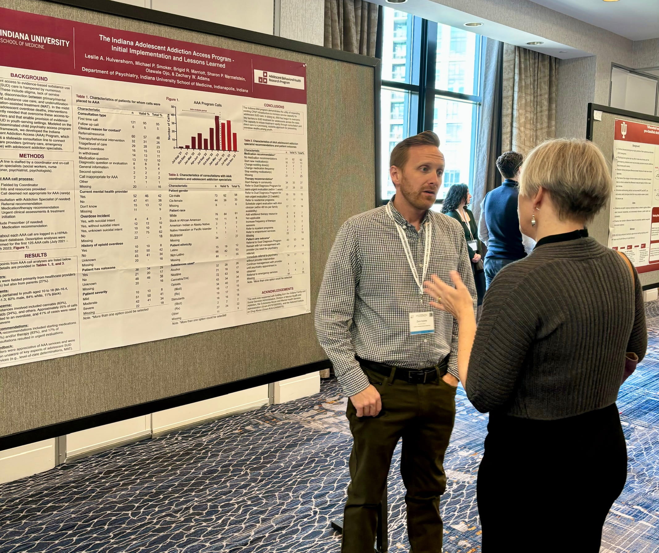 Dr. Zach Adams discussing with another person at a conference with poster for Behavioral Health behind him