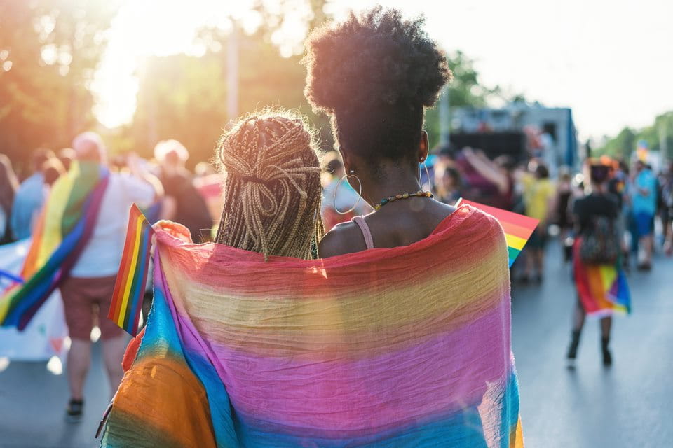 two black women stand wrapped in a pride flag looking into the sunshine