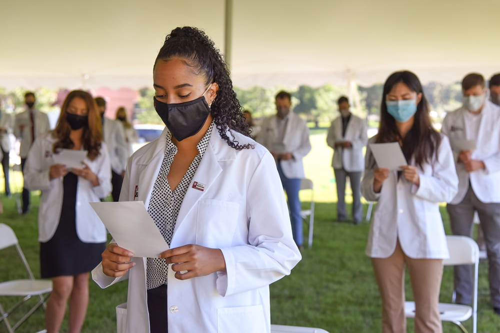 medical students in white coats and masks stand in proper social distancing while taking the physician's oath