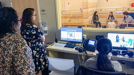 Three women watch a simulation from behind a women. In the room are a male physician and two young women.