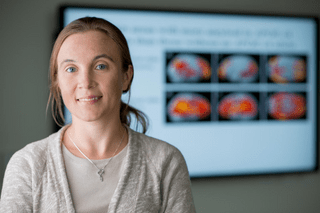 Researcher, Shannon Risacher stands in the foreground smiling with arms crossed. In the background is a screen with a blurred image that appears to reflect brain scans.