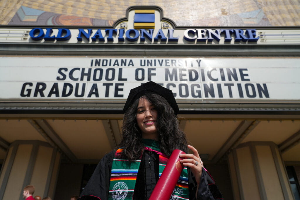 a student in graduation robes and a stole recognizing their latina cultural heritage stands in front of the Old National center holding their diploma. The marquee on the theater reads" IU School of Medicine Graduate Recognition"