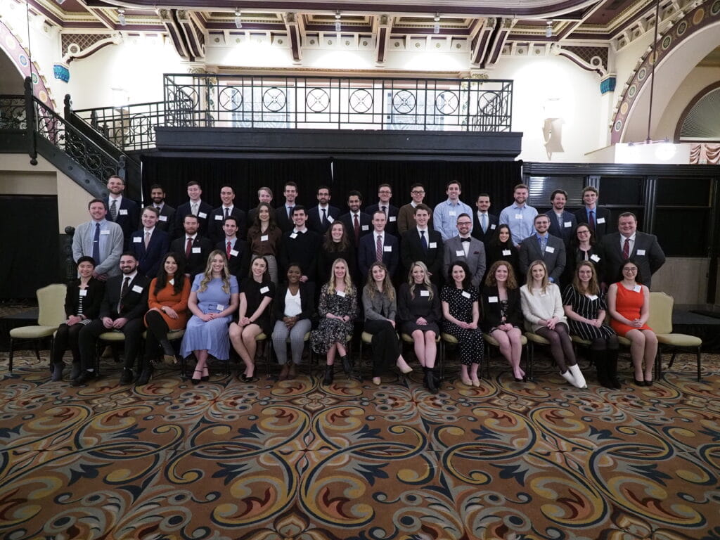3 rows of sharply-dressed students with name tags, all smiling in a fancy event center