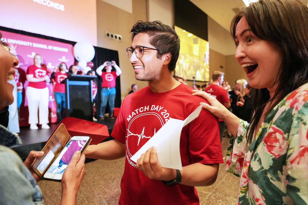 a student smiles with his match day envelope in hand while an astonished family member clasps his shoulder