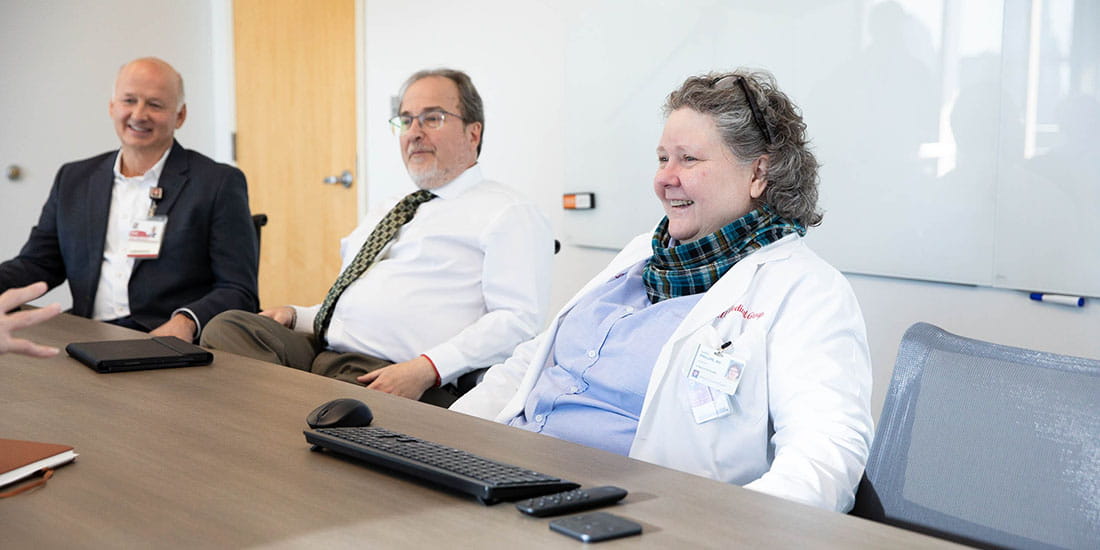 A group of professionals sitting at a conference table, including two men in business attire and a woman in a white lab coat and scarf, engaged in a meeting and smiling.