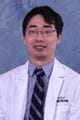 Cyrus wearing a lab coat and tie. He has dark short hair and glasses.