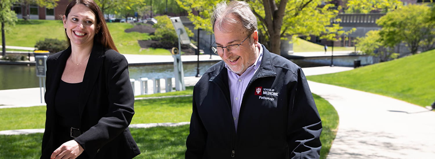 Two colleagues share a moment of laughter as they walk through a scenic campus path, surrounded by vibrant green spaces. Their friendly rapport reflects the collaborative and welcoming environment of the institution.