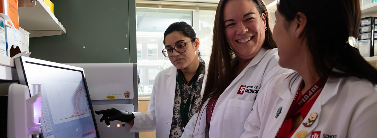 Aerial view of Indiana University campus buildings surrounding a peaceful, circular pond with a stepped amphitheater along its edge. Modern buildings with large glass windows are bathed in the warm glow of sunrise or sunset, creating a tranquil and inviting atmosphere. Trees and landscaped pathways enhance the scenic, open-campus layout.