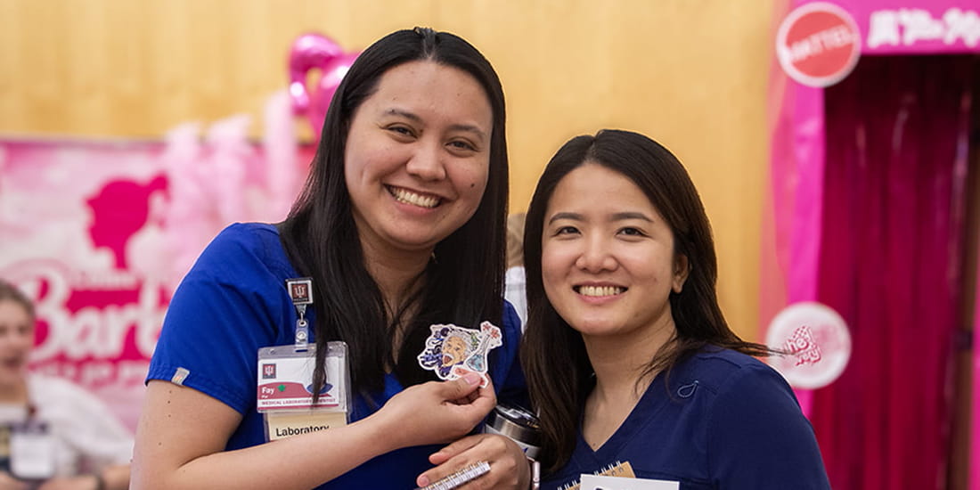 Two women wearing blue medical scrubs, smiling as they pose for a photo at a medical-themed event. One woman is holding up a sticker featuring an illustrated face, while the other holds a tumbler.