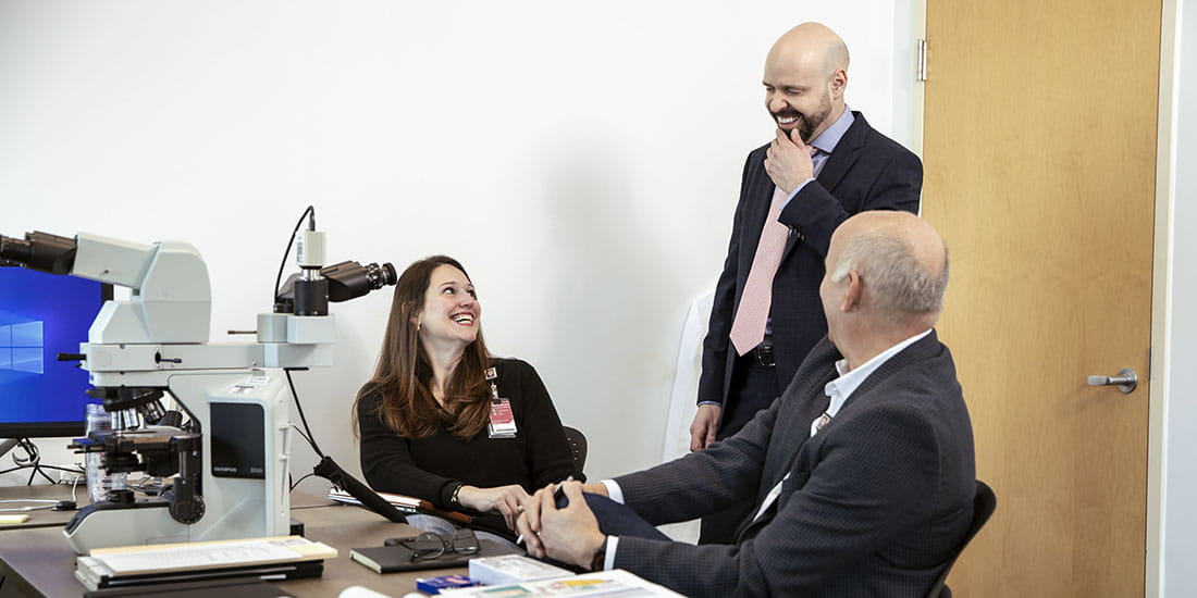 A meeting between colleagues, including a man in a suit and tie and two other professionals, one seated at a microscope, all smiling and engaged in conversation in an office setting.